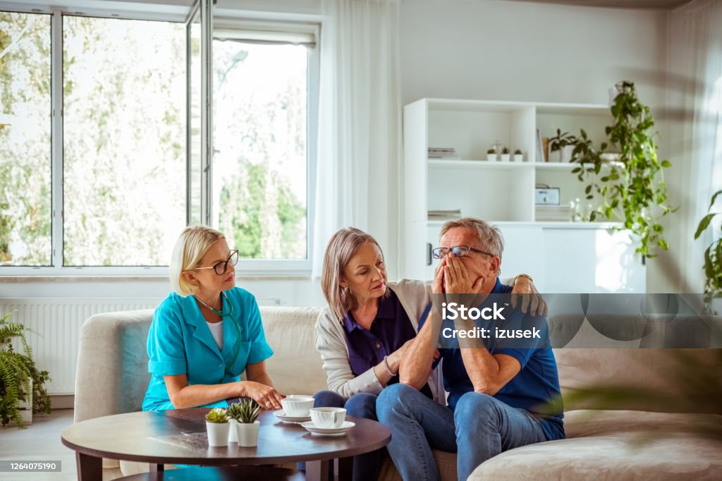 Nurse talking with worried senior man in hospital waiting room Worried senior man feeling unwell, getting bad news. Senior woman consoling him. They are sitting with nurse in hospital waiting room. Cancer - Illness Stock Photo