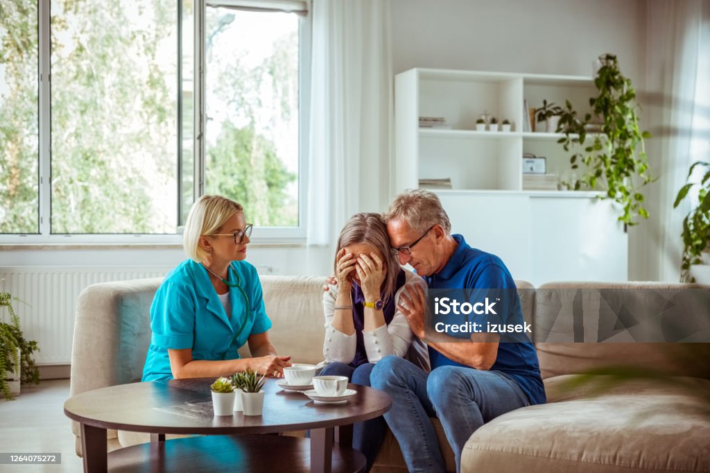 Nurse talking with worried senior woman Worried senior woman feeling unwell, getting bad news. Senior man consoling her. They are sitting with nurse in hospital waiting room. Sadness Stock Photo
