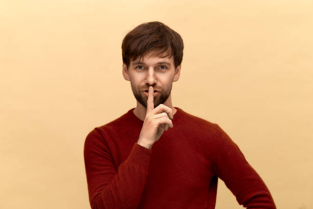 istock Keep silence. Photo of young man with beard wearing sweater, keeps fore finger over lips, makes silence gesture, posing against yellow background. 1264077250