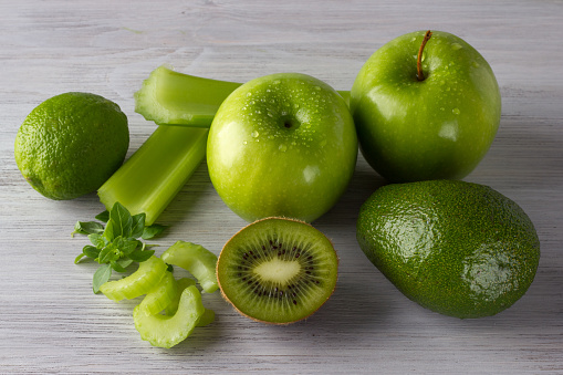 Fresh green fruits and vegetables: apples, lime, kiwi, basil, avocado, celery on the white wooden table