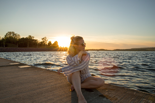 A young blonde woman lies on a jetty by a lake. The sun sets in the background. The warmth is within reach. The sun's rays can be seen. The waves are crashing.
