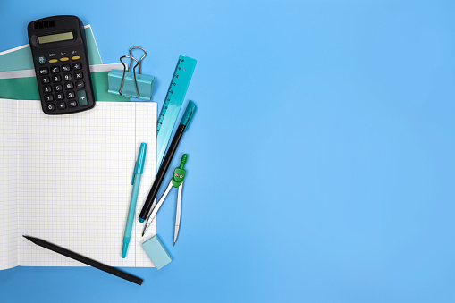 Math lessons flat lay blue banner. Back to school concept. Calculator, ruler, compasses, pen, pencil, eraser on white checkered open notebook. Squared sheet of a copybook. Blank page copy space.