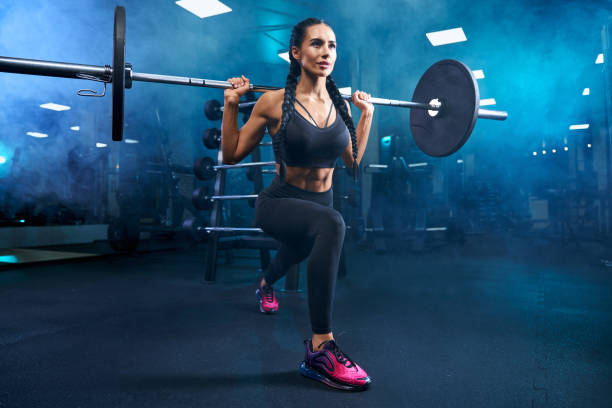 Fitnessoman practicing lunges using barbell in gym. Gorgeous brunette female with long braids doing squats using barbell. Side view of srtong concentrated fitnesswoman with perfect muscular body training legs in gym in dark atmosphere and smoke. body building stock pictures, royalty-free photos & images