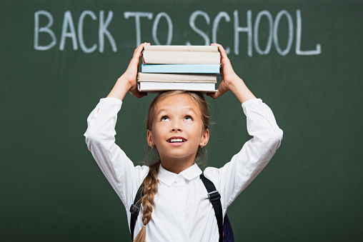 selective focus of smiling schoolgirl holding stack of books above head near chalkboard with back to school text