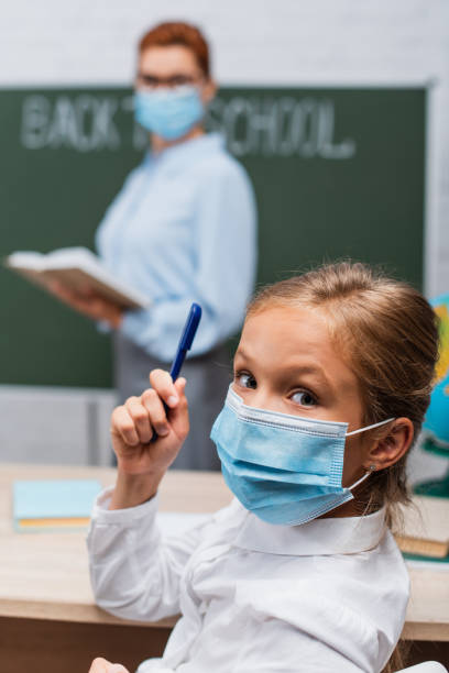 selective focus of schoolgirl in medical mask holding pen and looking at camera, and teacher with book near chalkboard selective focus of schoolgirl in medical mask holding pen and looking at camera, and teacher with book near chalkboard teacher classroom child education stock pictures, royalty-free photos & images