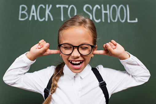 selective focus of displeased schoolgirl in eyeglasses plugging ears with fingers near back to school inscription on chalkboard