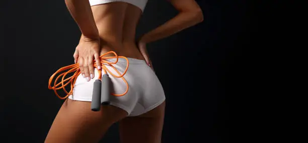 Slim sporty girl showing her taned body back in white underwear posing close-up with orange jumping-rope with the black background text space to the right