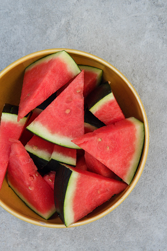 Yellow bowl with cut slices of watermelon, a half of a seedless watermelon and a knife with wooden handle on the concrete background