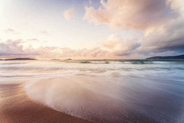 This photograph captures the magnificent scenery of the coast at dusk. The sky is adorned with golden clouds, with sunlight filtering through, casting a soft glow over the ocean waves. The interplay of light and shadow on the water creates a mesmerizing effect. In the distance, a lone lighthouse stands tall on the rocky shore, quietly watching over the sea, adding a sense of solitude and steadfastness. This image is perfect for themes related to nature, the sea, tranquility, and peaceful moments.