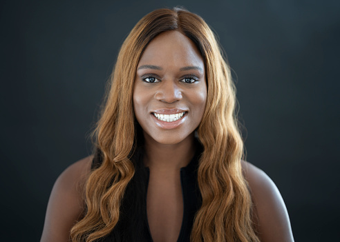 Front view of 26 year old woman with long wavy brown hair wearing sleeveless black top and smiling at camera against black background.