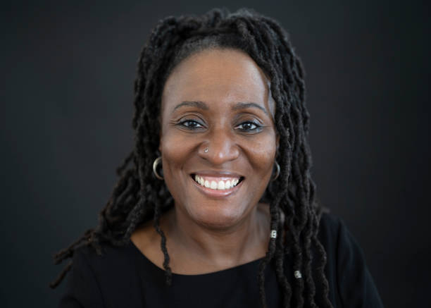 Front view portrait of cheerful mature black woman Headshot of 48 year old woman with long cornrow braids wearing hoop earrings, dark top with round neckline, and smiling at camera against black background. black hair braiding stock pictures, royalty-free photos & images