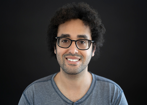 Front view portrait of 28 year old man with short hairstyle and facial hair stubble wearing gray Henley t-shirt and smiling at camera against black background.