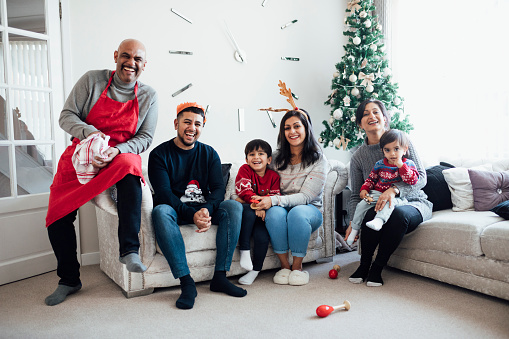 A full-length portrait of a multi-generation family sitting on sofas in their home living room during the Christmas period time. They are sitting side by side while looking at the camera and smiling.