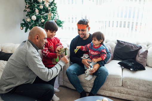 A multi-generation family sitting in the home living room during the Christmas period time. The children are playing with dinosaurs while the father and grandfather watch them.