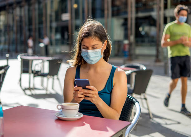 istock COVID-19 outbreak. Young woman wearing protective surgical face mask and using mobile phone in a coffee shop terrace in urban summer. Coronavirus, the New Normal and mandatory use of face mask 1264115219