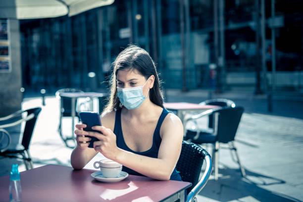 istock COVID-19 outbreak. Young woman wearing protective surgical face mask and using mobile phone in a coffee shop terrace in urban summer. Coronavirus, the New Normal and mandatory use of face mask 1264115291