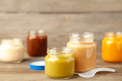 Glass jars with nutrient baby food on grey background. Vegetable puree with spoon.