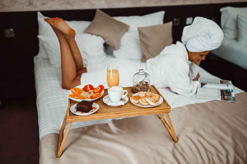 Beautiful young woman wearing a bathrobe lying down in bed, eating breakfast and reading a magazine