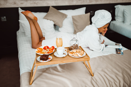 Beautiful young woman wearing a bathrobe lying down in bed, eating breakfast and reading a magazine