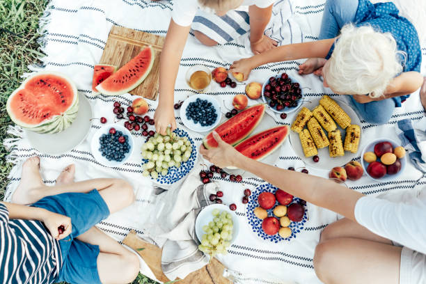 Top view of the hands of people taking food. Fresh summer vitamin harvest. Top view of a picnic tablecloth filled with plates of berries and fruits, watermelon and corn. Top view of the hands of people taking food. Fresh summer vitamin harvest. Friendly community. cottagecore stock pictures, royalty-free photos & images