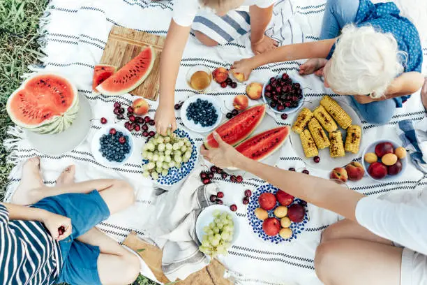 Top view of a picnic tablecloth filled with plates of berries and fruits, watermelon and corn. Top view of the hands of people taking food. Fresh summer vitamin harvest. Friendly community.
