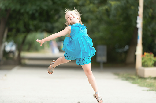 small baby girl or cute happy child with adorable smiling face and bow in blonde hair in blue dress jumping in summer outdoor on blurred background.