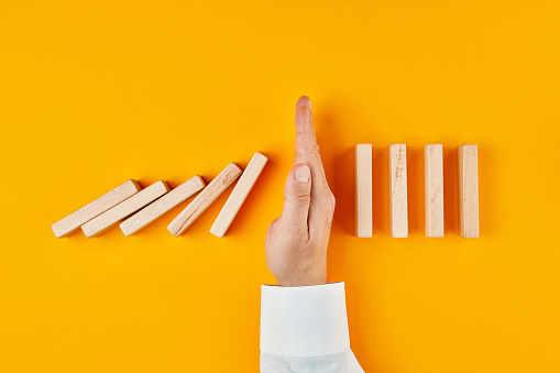 Hand of a businessman stopping domino effect on yellow background. Concept of risk protection, business solution or successful intervention strategy.