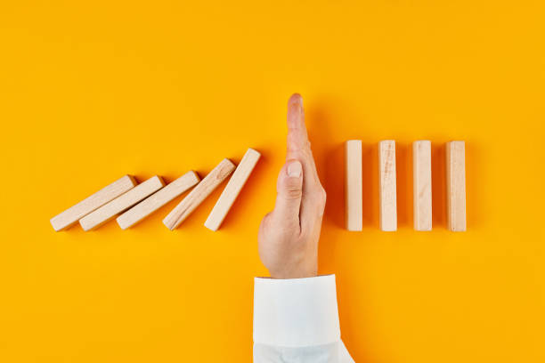 Hand of a businessman stopping domino effect on yellow background Hand of a businessman stopping domino effect on yellow background. Concept of risk protection, business solution or successful intervention strategy. hand stopping dominos stock pictures, royalty-free photos & images