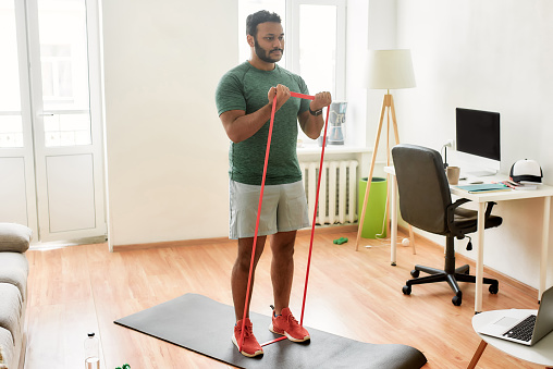 Tone up your body. Full length shot of young active man watching online video training on laptop, exercising with resistance band during morning workout at home. Sport, healthy lifestyle
