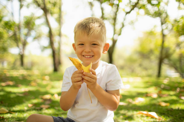 Happy little blonde boy playing with the leaves in the park.