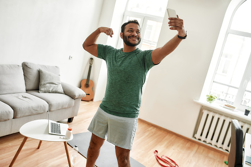 Be physically fit. Excited bearded active man holding smartphone, showing biceps while taking a selfie during morning workout at home. Fitness, motivation concept. Horizontal shot