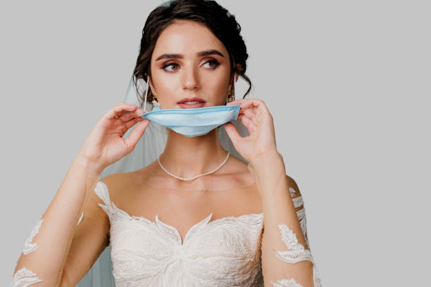 bride in medical mask and wedding bouquet at coronavirus covid-19 quarantine period. attractive girl takes off mask and looks in camera in studio on white background - hairstyle love wedding photography imagens e fotografias de stock