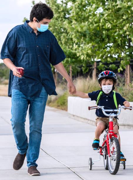 istock young man walking his son back to school, boy is riding his bicycle both wearing protective face masks 1264156937