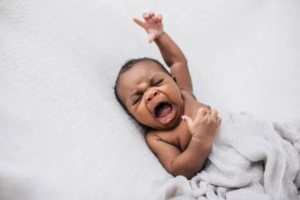 Photo of crying and upset and angry African-American newborn baby boy lying on a cream blanket