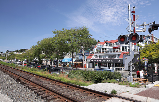 White Rock, Canada - August 1, 2020: Pedestrians wait near the train tracks and a level crossing on scenic Marine Drive. Upscale residences and popular businesses face Semiahmoo Bay in Metro Vancouver.