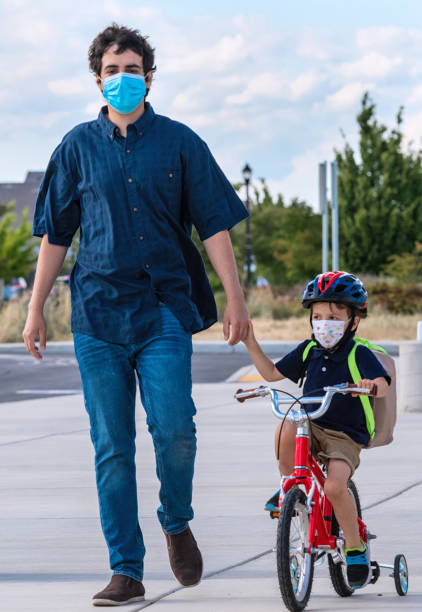 istock young man walking his son back to school, boy is riding his bicycle both wearing protective face masks 1264159155