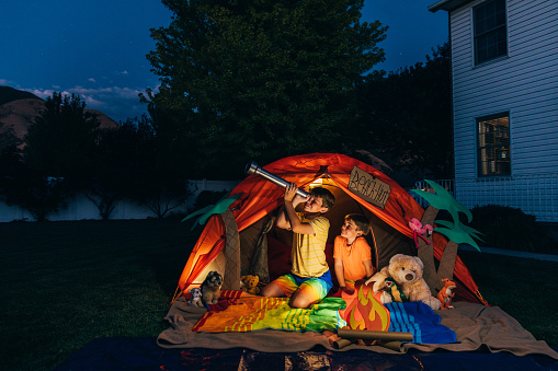 Two brothers camp out in the backyard of their home due to the coronavirus restrictions and quarantine. The boys have pitched a tent and created a beach scene with stuffed animal friends and have a fake campfire. They are searching the twilight for the stars in the sky.