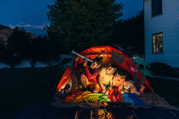 Backyard Staycation and Exploration A dad with his two sons camp out in the backyard of their home due to the coronavirus restrictions and quarantine. They have pitched a tent and created a beach scene with stuffed animal friends and have a fake campfire. They are searching the twilight for the stars in the sky. staycation stock pictures, royalty-free photos & images