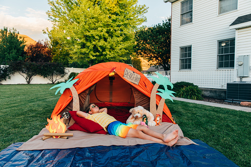 A dad with his son and young daughter camp out in the backyard of their home due to the coronavirus restrictions and quarantine. They have pitched a tent and created a beach scene with stuffed animal friends. They are making the best of their situation and long to return to the real outdoors.