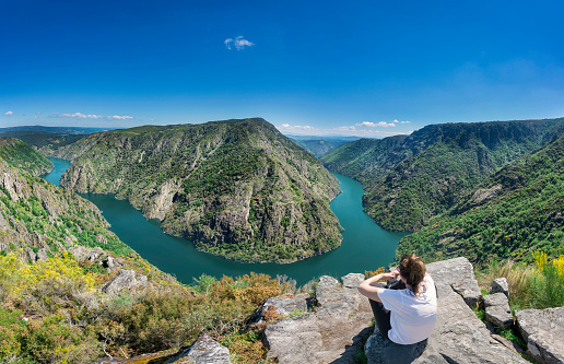A woman contemplates the incredible Sil Canyon in the Ribeira Sacra from a fabulous viewpoint where she can see a meander of the Sil river. The woman is sitting in a rock enjoying the impressive landscape