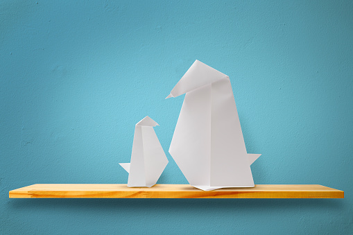 Blank origami penguin parent and child on a wooden shelf against blue wall.