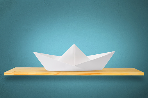 Blank origami paper boat on a wooden shelf against blue wall.