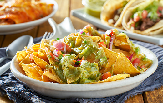 Stock photo showing close-up, elevated view of homemade loaded nachos recipe. This snack contains fresh, crunchy tortilla chips covered in melted cheese with cherry tomatoes, black olives and Jalapeno peppers on an oval serving dish.
