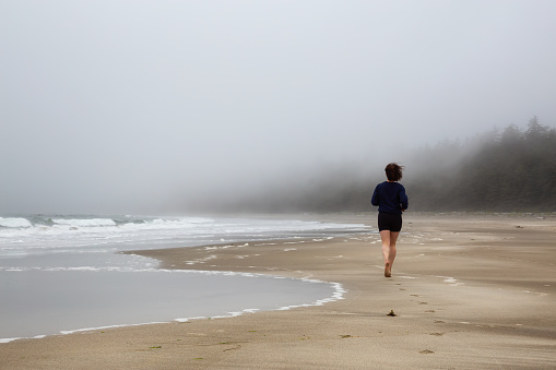 Athletic Caucasian Girl Running on a Sandy Beach near Pacific Ocean Coast during a cloudy and foggy morning. Taken at Raft Cove, Vancouver Island, British Columbia, Canada.