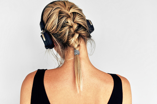 Horizontal rear view image of a beautiful blonde young woman listening to music with headphones. Back view portrait of pretty female wearing a black dress listens to the audiobook in headphones.