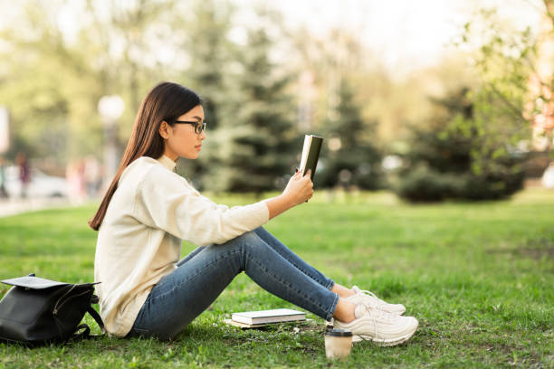 Girl reading a book sitting in the park, copyspace Learning Concept. Side view full length portrait of focused asian teen reading a book sitting on grass in parkland, copyspace hot filipina women stock pictures, royalty-free photos & images