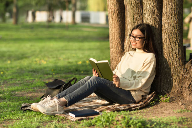 Girl reading a book sitting under the tree Side view of pleased brunette asian woman in eyeglasses sitting under the tree and reading book in park hot filipina women stock pictures, royalty-free photos & images