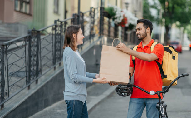 istock Shopping from home. Friendly courier with backpack and bicycle gives paper bag to client 1264231869