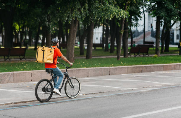 istock Courier bicycle delivery service. Man with large yellow backpack ride along road in park 1264231946