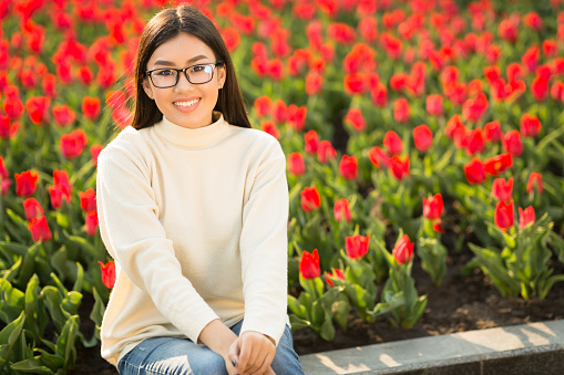 Spending Free Time Outdoors. Smiling asian teen wearing glasses, sitting on parapet near tulip garden, free space